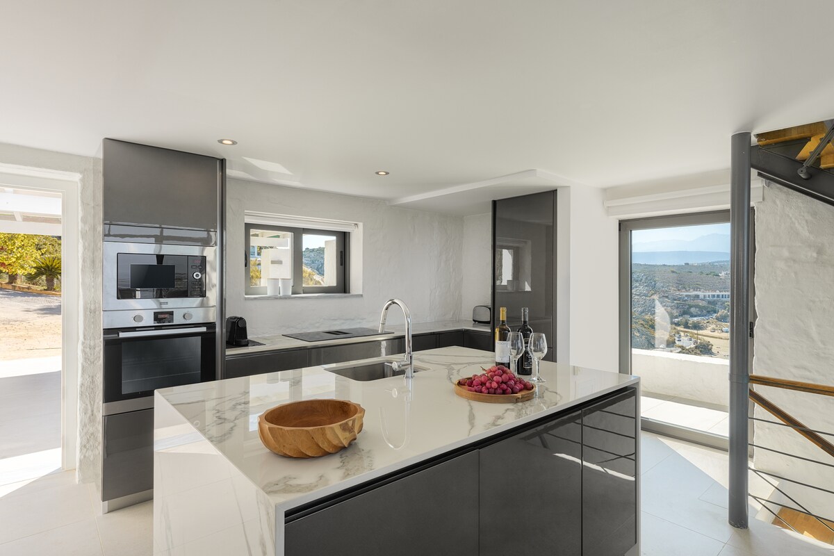 The kitchen features a modern design with sleek black cabinetry and a spacious island. Natural light flows in through large windows, offering views of the landscape. Equipped with high-end appliances, it is both functional and visually appealing, providing a comfortable setting for meal preparation.