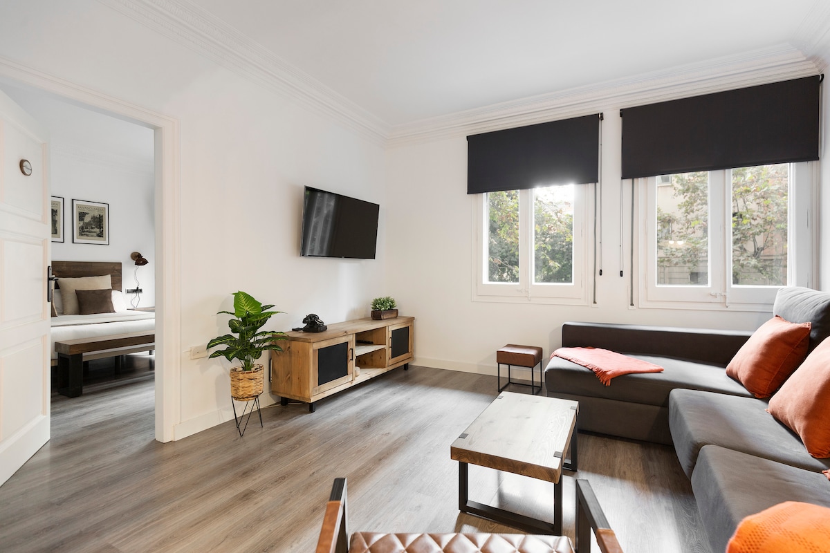 The living area features a comfortable sectional sofa adorned with orange pillows, complemented by a wooden coffee table. A wall-mounted TV is visible, and natural light streams in through two windows, which are dressed with simple black shades. A potted plant adds a touch of greenery.