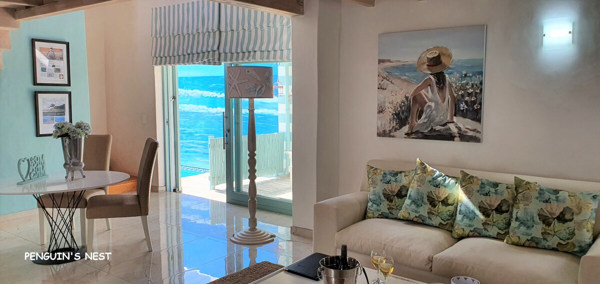 An inviting living area features a comfortable sofa adorned with floral cushions, along with a dining table for two. Large glass doors allow natural light to enter, revealing stunning ocean views. Artwork depicting a serene beach scene complements the decor.