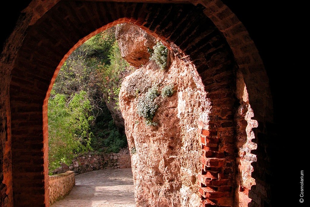 An arched stone doorway frames a pathway leading into a lush green area. The natural rock formations, adorned with patches of moss, provide a captivating contrast to the rustic brickwork of the archway.