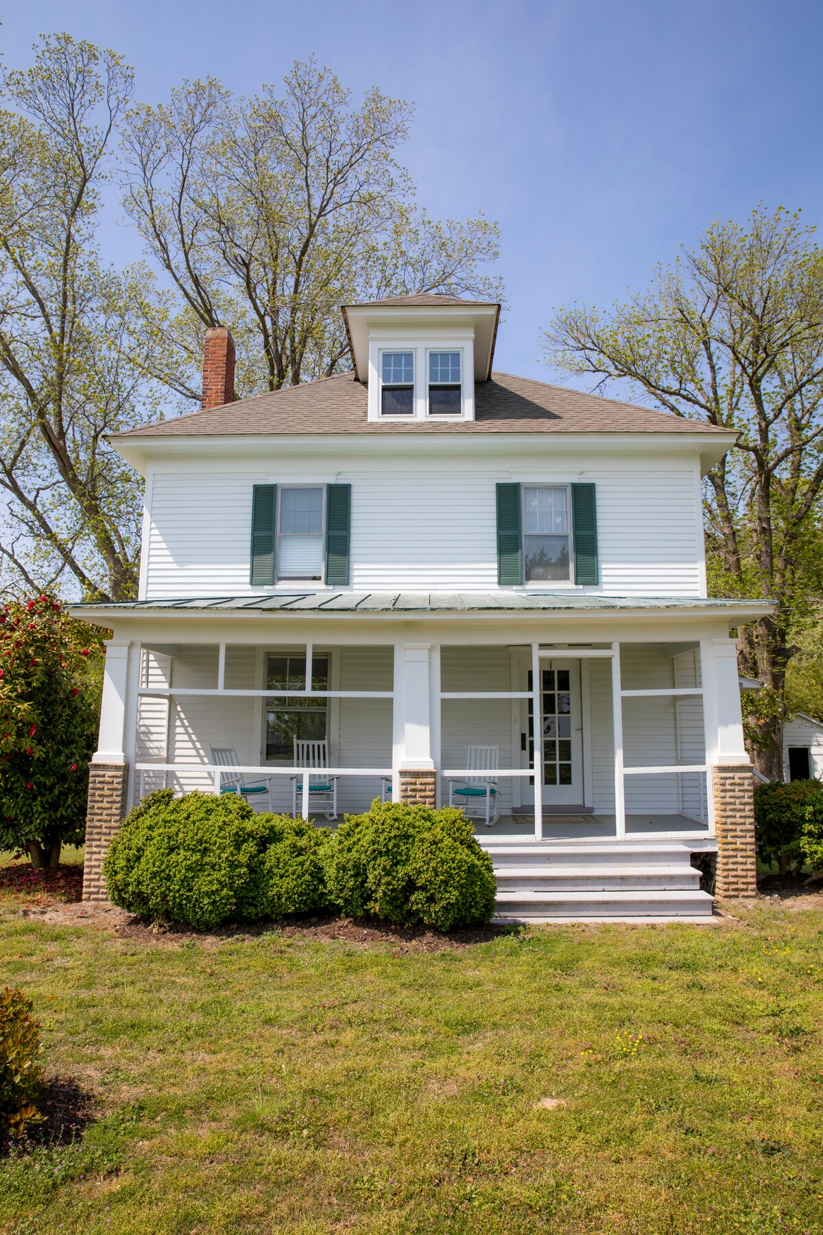 A classic two-story farmhouse is framed by lush greenery and tall trees. A welcoming porch with white rocking chairs offers a place to relax, while the gabled roof adds architectural interest. The exterior features crisp white siding with dark green shutters.