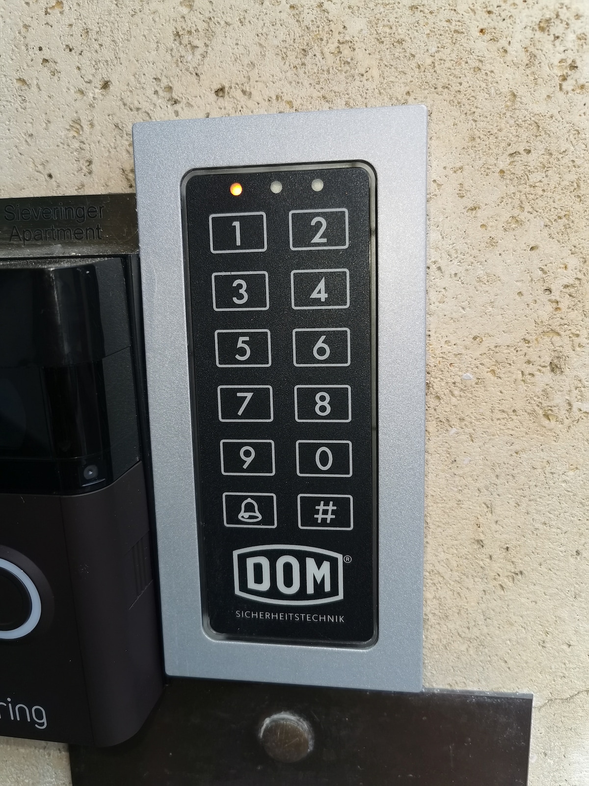 A keypad entry system is mounted on a textured wall, featuring a grid of numbers from 0 to 9. A small light is illuminated above the first button, indicating power or readiness. The keypad is labeled with the brand name 'DOM' and a privacy symbol.