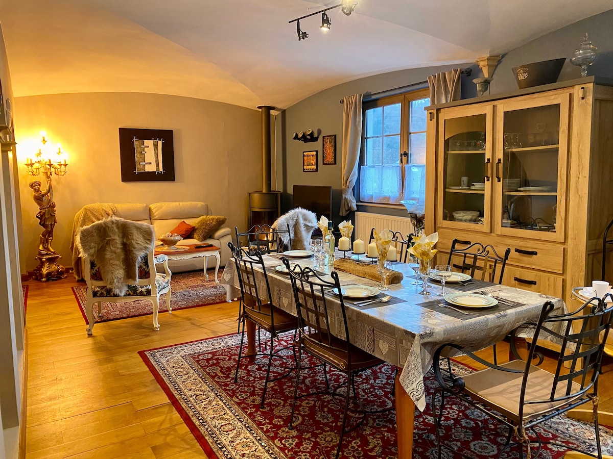 A spacious dining area features a long table set for several guests, accompanied by elegant chairs. A cozy seating area is present, with a sofa adorned with soft throws. Warm lighting creates a relaxing ambiance, while large windows allow natural light to filter in.