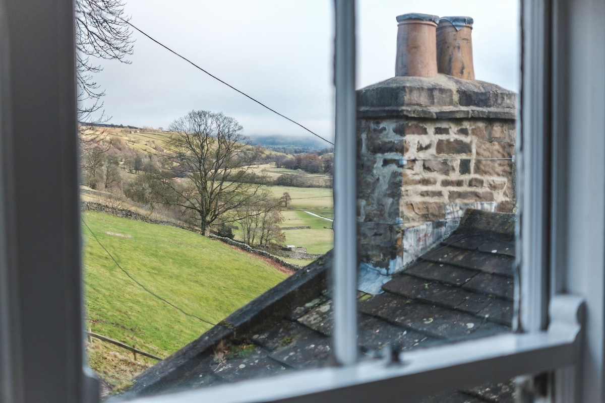 A view from the window reveals a serene landscape, featuring rolling green hills and distant trees under a cloudy sky. The stone chimney and slate roof are visible in the foreground, blending harmoniously with the tranquil scenery.