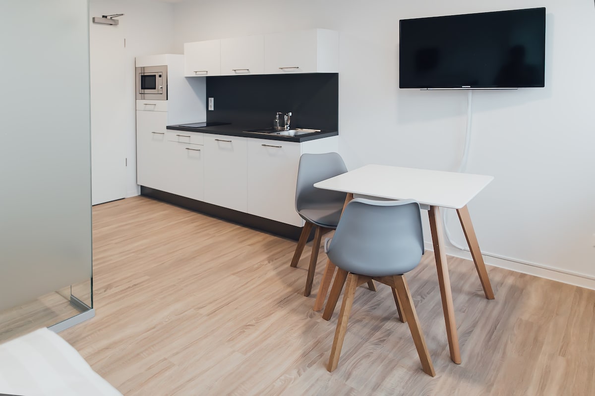 A modern kitchenette is featured with sleek cabinetry and a counter space that includes a sink and a microwave. A dining area with a white table and two grey chairs is visible, alongside a wall-mounted flat-screen TV. Light wood flooring enhances the spacious feel.