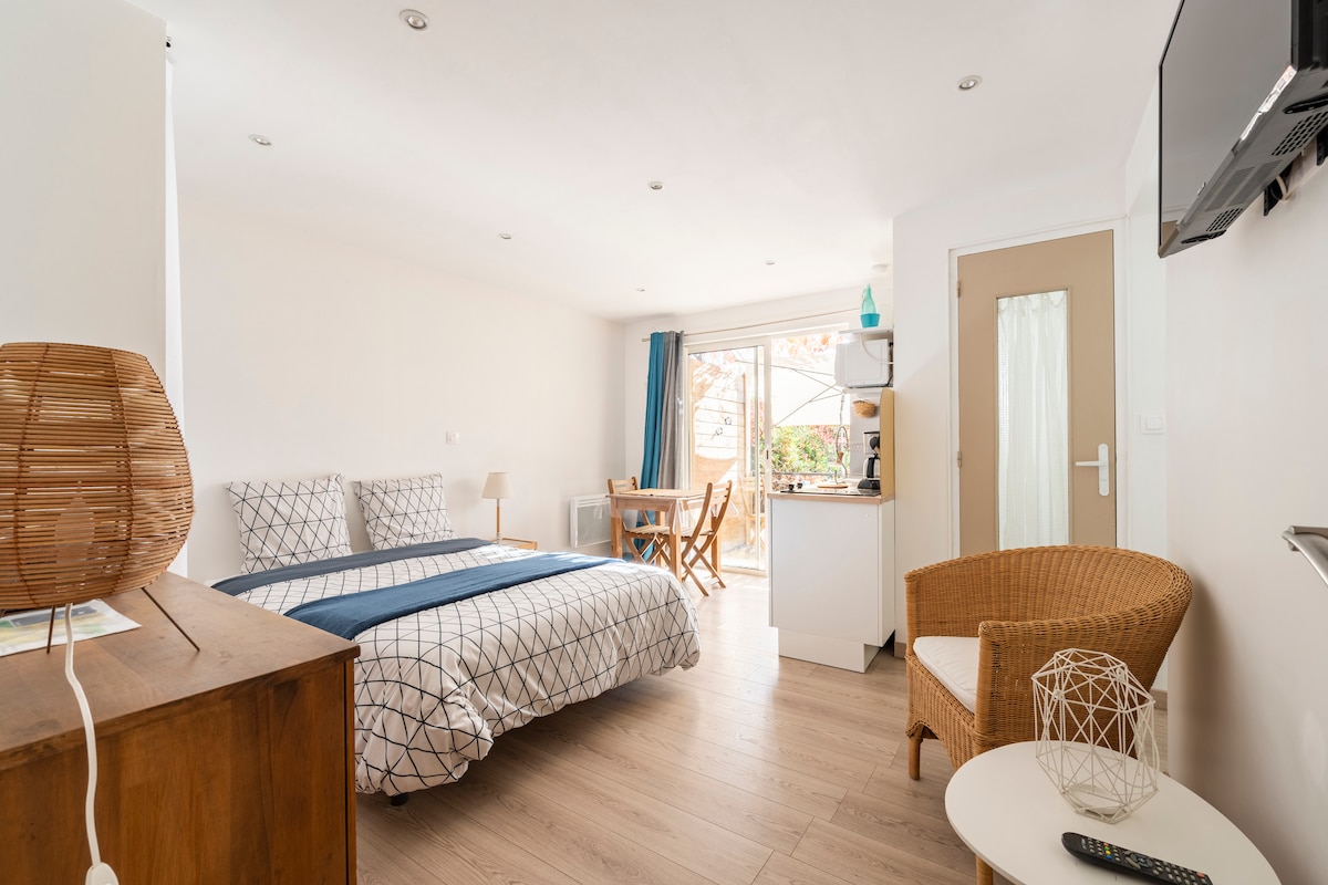 A bright studio features a double bed dressed in a geometric-patterned duvet. A small dining area with a wooden table and chairs is positioned beside a well-equipped kitchenette. Natural light enters through a window with light blue curtains, complementing the modern decor.