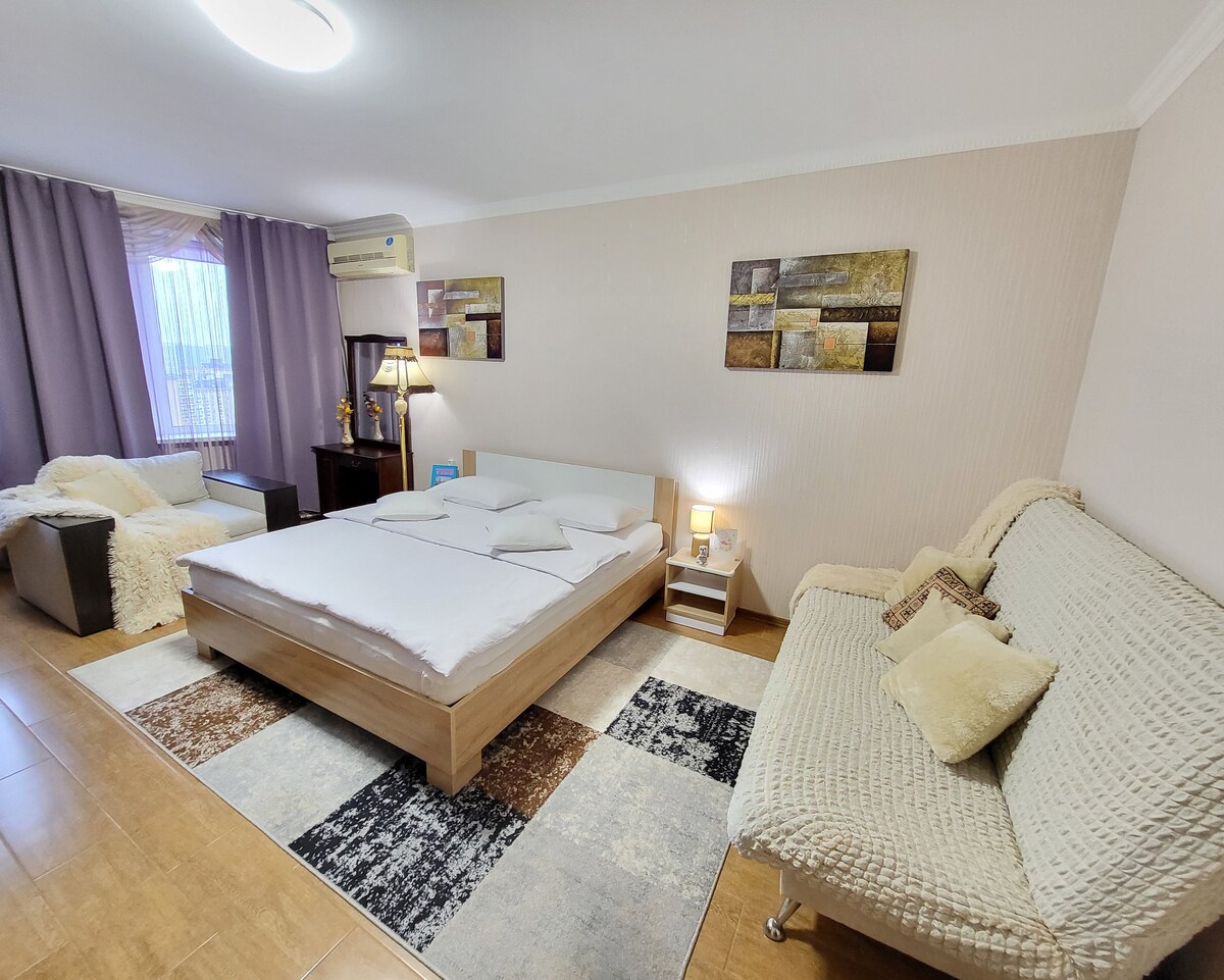 A spacious room features a modern wooden bed with soft bedding, complemented by a light-colored textured sofa and armchair. A small bedside table and lamp are positioned near the bed, while large windows allow natural light to fill the space.