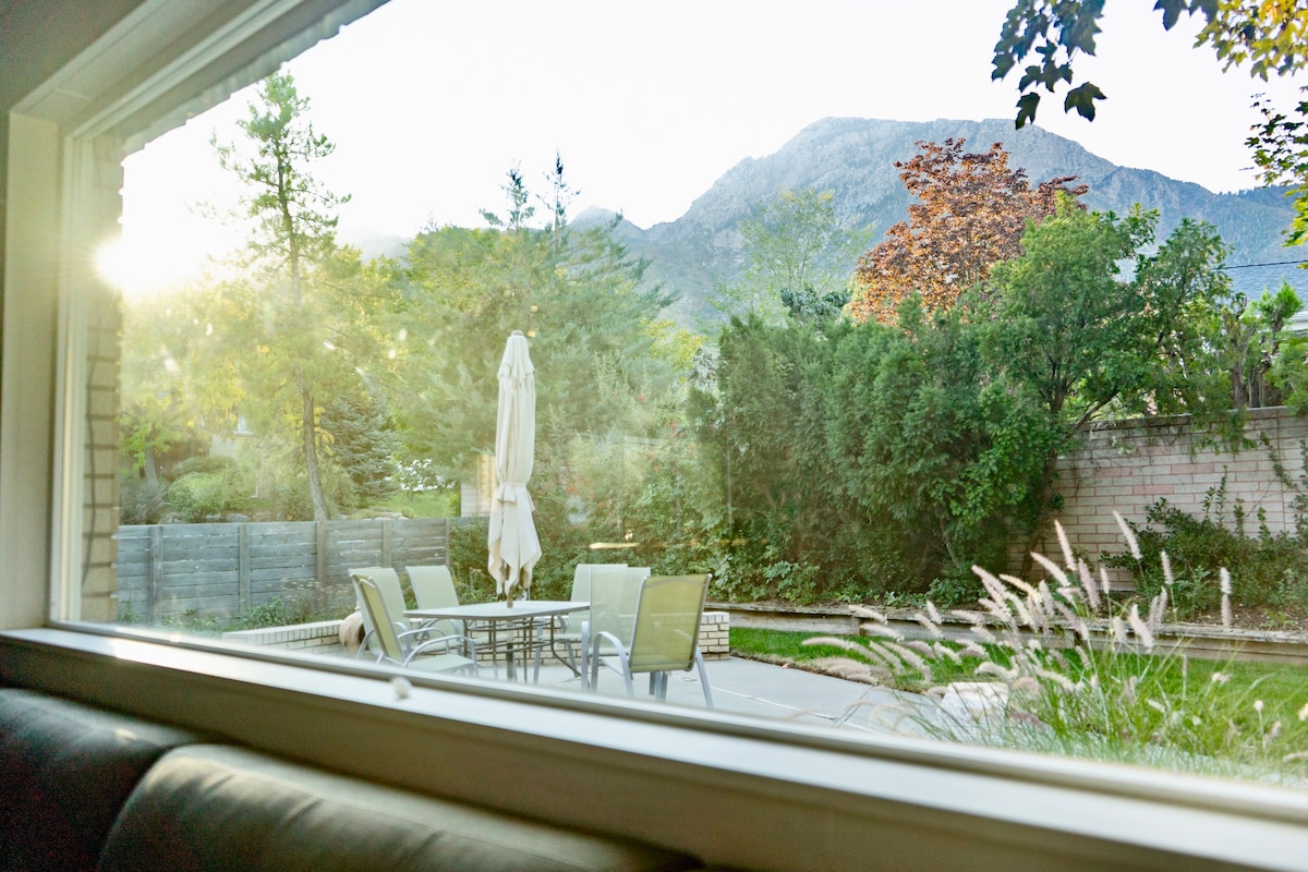 The view from a window showcases a serene outdoor space featuring a dining table with chairs, an umbrella, and landscaped greenery. The backdrop includes a mountain silhouette, while sunlight filters through the trees, creating a peaceful ambiance in the yard.
