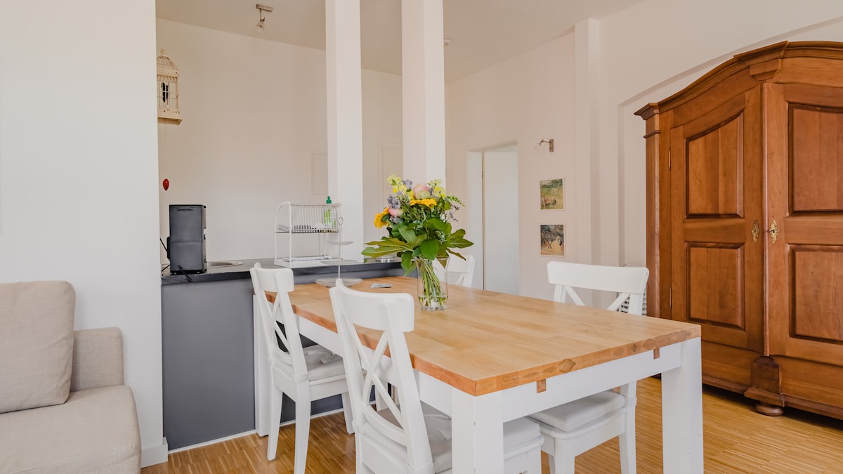 An airy dining area features a wooden table surrounded by white chairs. A bouquet of flowers rests in the center. In the background, a dark kitchen counter is visible, along with a large wooden wardrobe. Soft lighting creates a welcoming atmosphere.