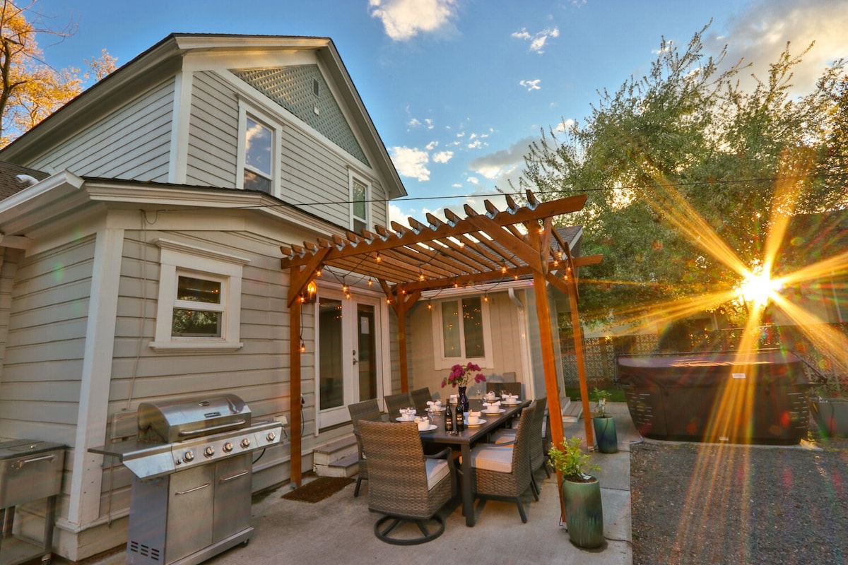 A private outdoor patio features a covered dining area with a table set for several guests. A BBQ grill is positioned nearby, accompanied by potted plants. Sunlight streams through the trees, creating a warm ambiance as the evening approaches.