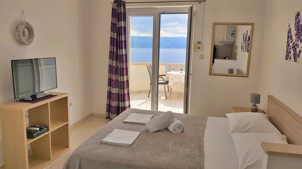 A bright room features a spacious bed covered with soft linens and neatly arranged towels. A TV is positioned on a wooden unit, while large doors open to a sea view balcony. Light flows in, creating a calm and relaxing atmosphere.