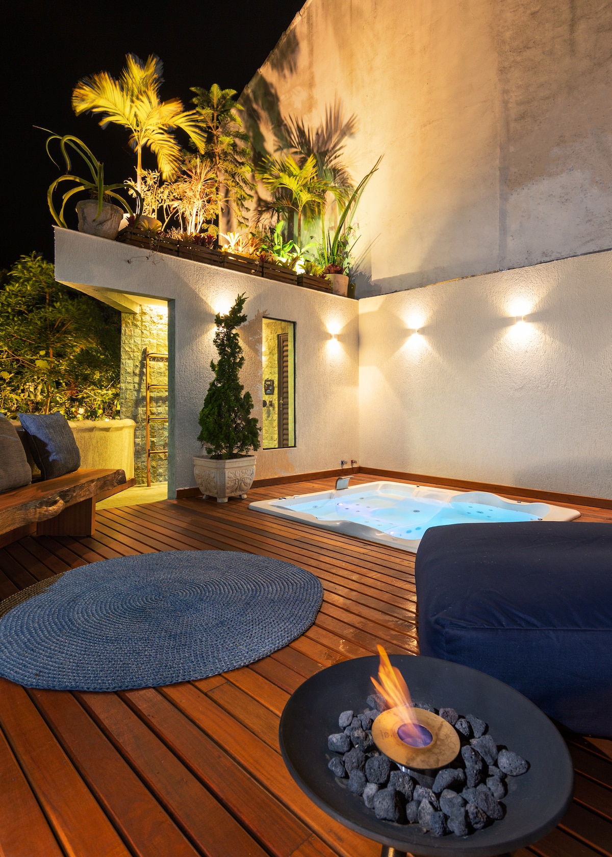 A serene spa area features a wooden deck, with a warm jacuzzi illuminated under soft lighting. Lush tropical plants are positioned around the space, and a fire pit sits nearby, providing a cozy focal point for relaxation.