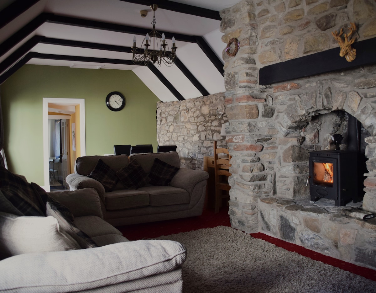 A cozy lounge area features a stone fireplace with a log burning stove, surrounded by exposed stone walls. Comfortable seating is arranged, and a dining area can be seen through an archway. Dark wooden beams span the ceiling, enhancing the rustic atmosphere.