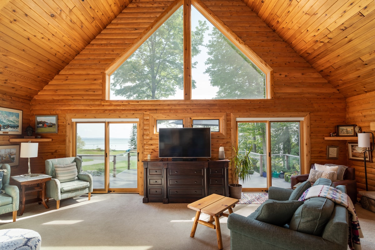 A spacious living area features vaulted wood ceilings and large windows, allowing natural light to illuminate the room. Comfortable seating options are arranged around a wooden coffee table, with a television mounted on a sturdy dresser. Scenic views of the outdoors are visible through the glass doors.