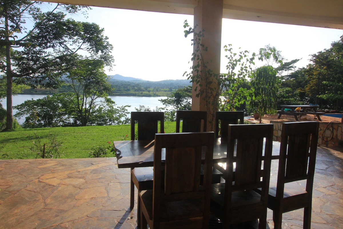 A dining table with six wooden chairs is positioned on a stone patio, overlooking a serene view of The River Nile. Lush greenery frames the scene, while distant hills provide a gentle backdrop under clear blue skies.