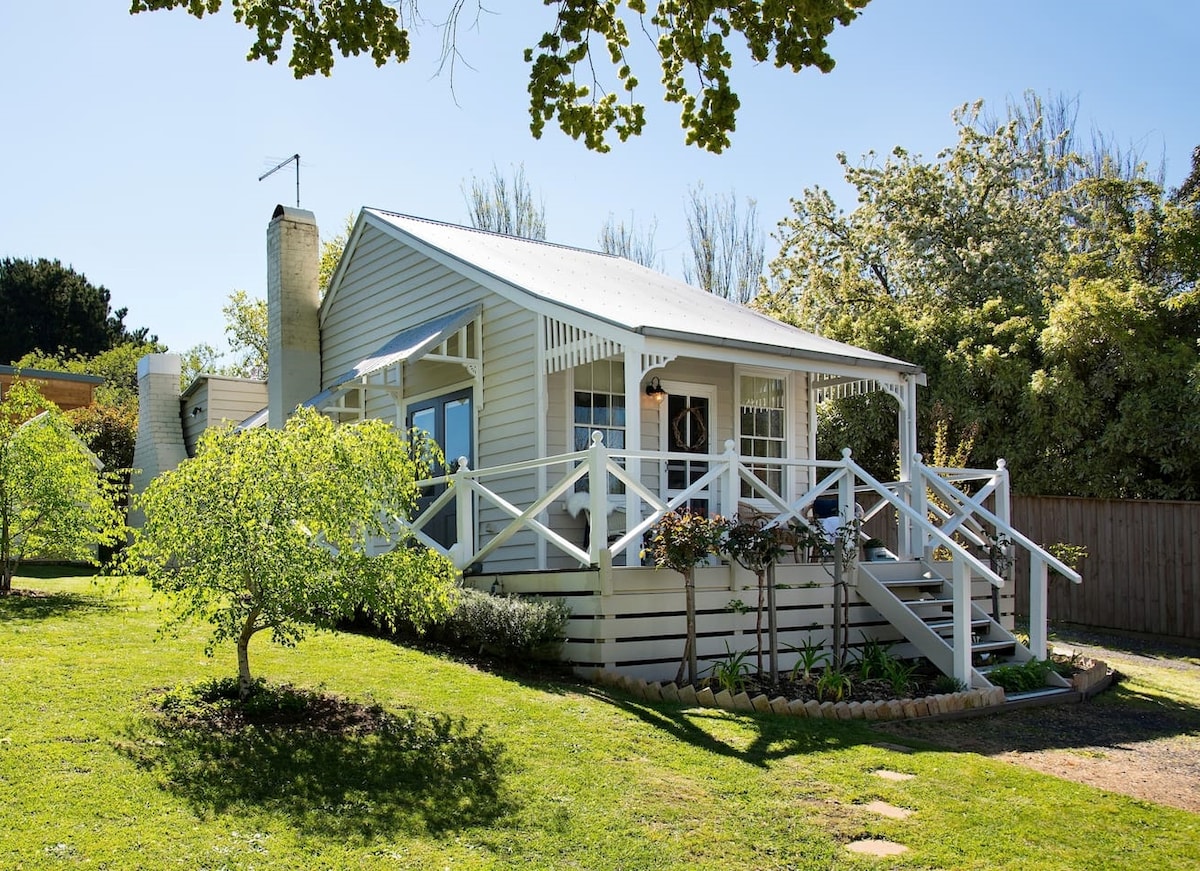 The charming cottage features a welcoming front porch with white railings, surrounded by a well-kept lawn and colorful garden. Large windows allow natural light to fill the space, while the roof peaks gently under a clear blue sky.