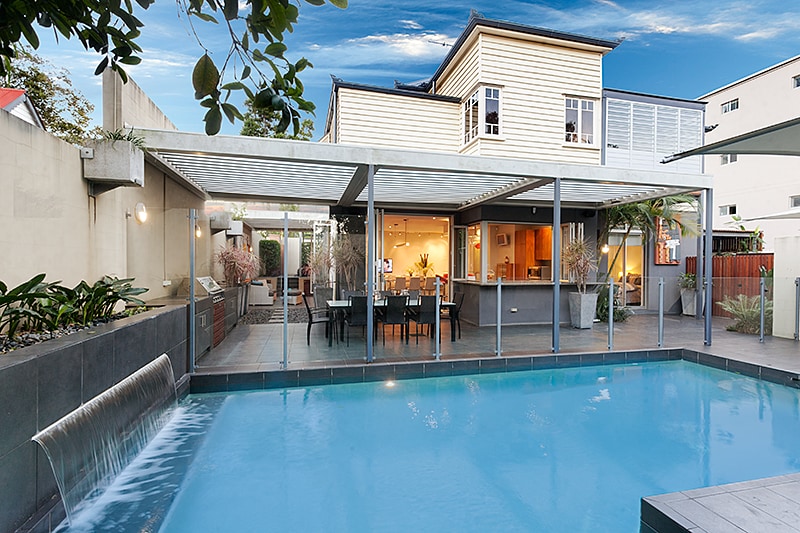 A private outdoor area features a sparkling pool surrounded by glass fencing. A covered patio area includes a dining table and chairs, with an outdoor kitchen space visible in the background. Lush greenery enhances the serene environment.