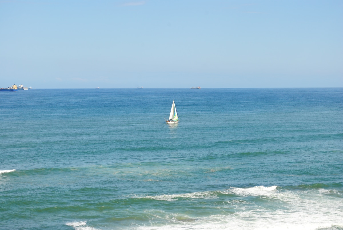A sailboat glides across a calm sea, surrounded by expansive blue waters. The horizon meets a clear sky, with a few vessels visible in the distance. Gentle waves break along the shore, creating a serene and tranquil scene.