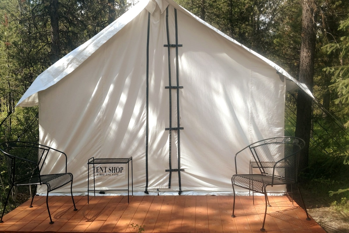 A canvas tent is situated on a wooden platform, featuring a solid front entrance secured with straps. Two outdoor chairs are positioned on the deck, providing a space to relax and enjoy the surrounding natural setting.
