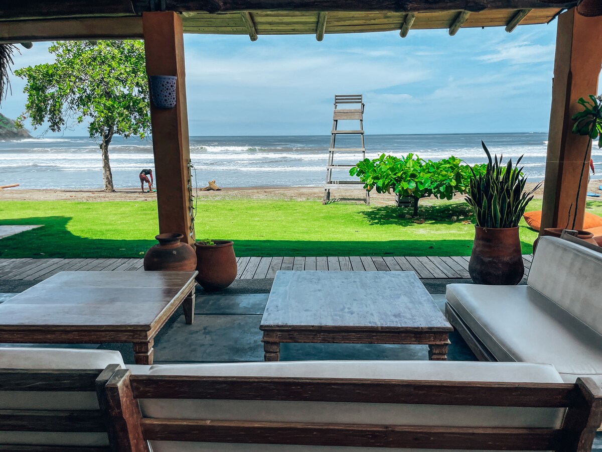 A shaded outdoor lounge area features wooden seating arrangements with white cushions and a large coffee table. Lush green grass extends toward the shore, where gentle ocean waves lap against the beach. A lifeguard tower stands in the distance, framed by palm trees.