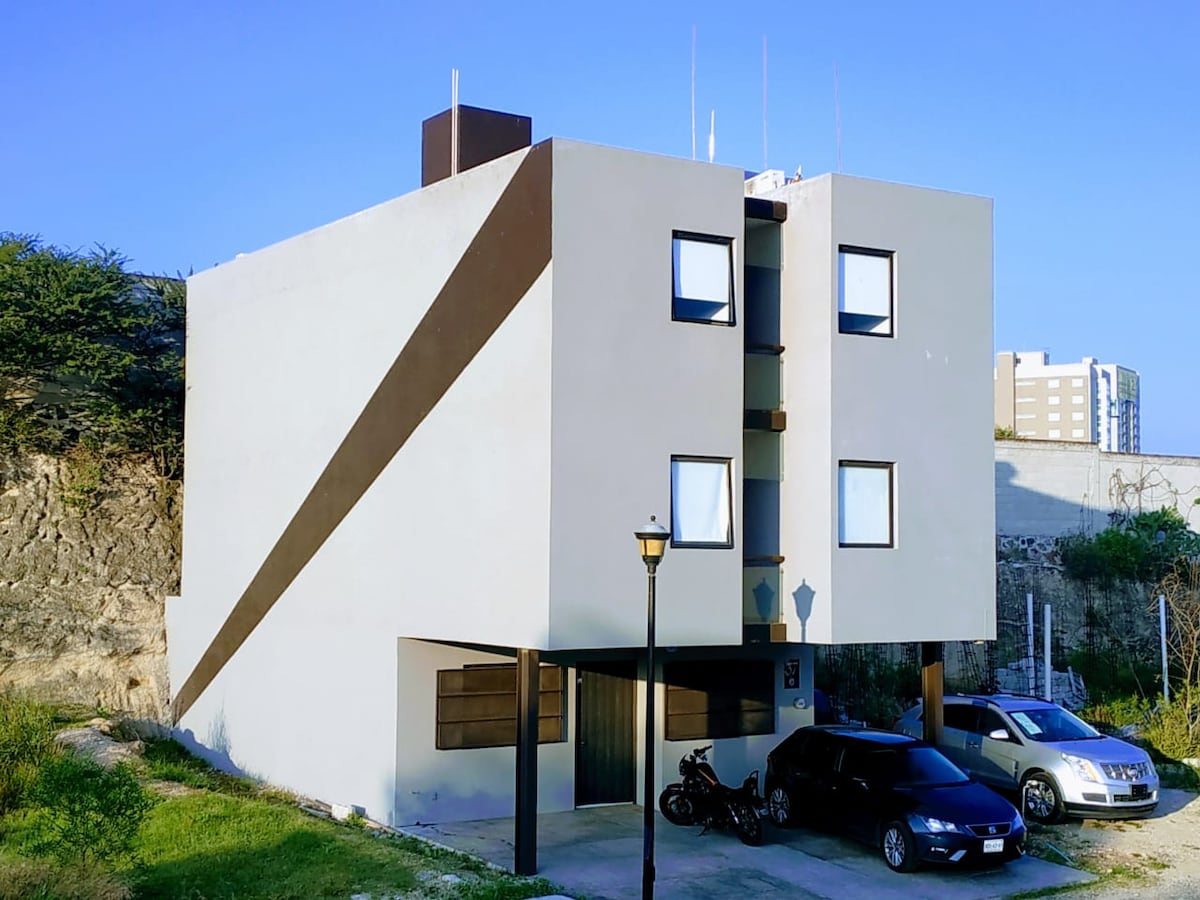 A modern three-story building features clean lines and a minimalist design. Large square windows provide ample natural light, while a distinctive brown accent stripe enhances the facade. The ground level includes outdoor parking spaces and is framed by lush greenery and distant city buildings.