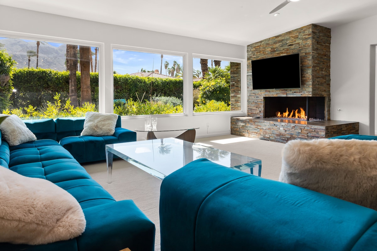 A spacious living room features a modern sectional sofa in teal and a glass coffee table. Large windows provide views of lush greenery and mountain scenery, while a stone fireplace with a mounted TV adds a cozy element to the room.