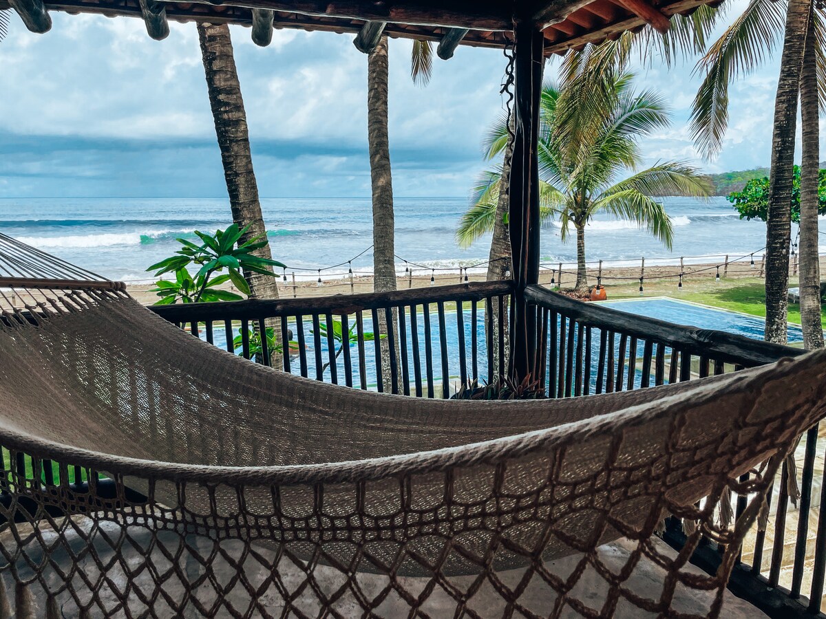 A hammock is positioned on a private balcony, allowing views of the ocean and lush palm trees. The sound of waves can be heard in the background, and a tranquil pool is visible beneath cloudy skies.