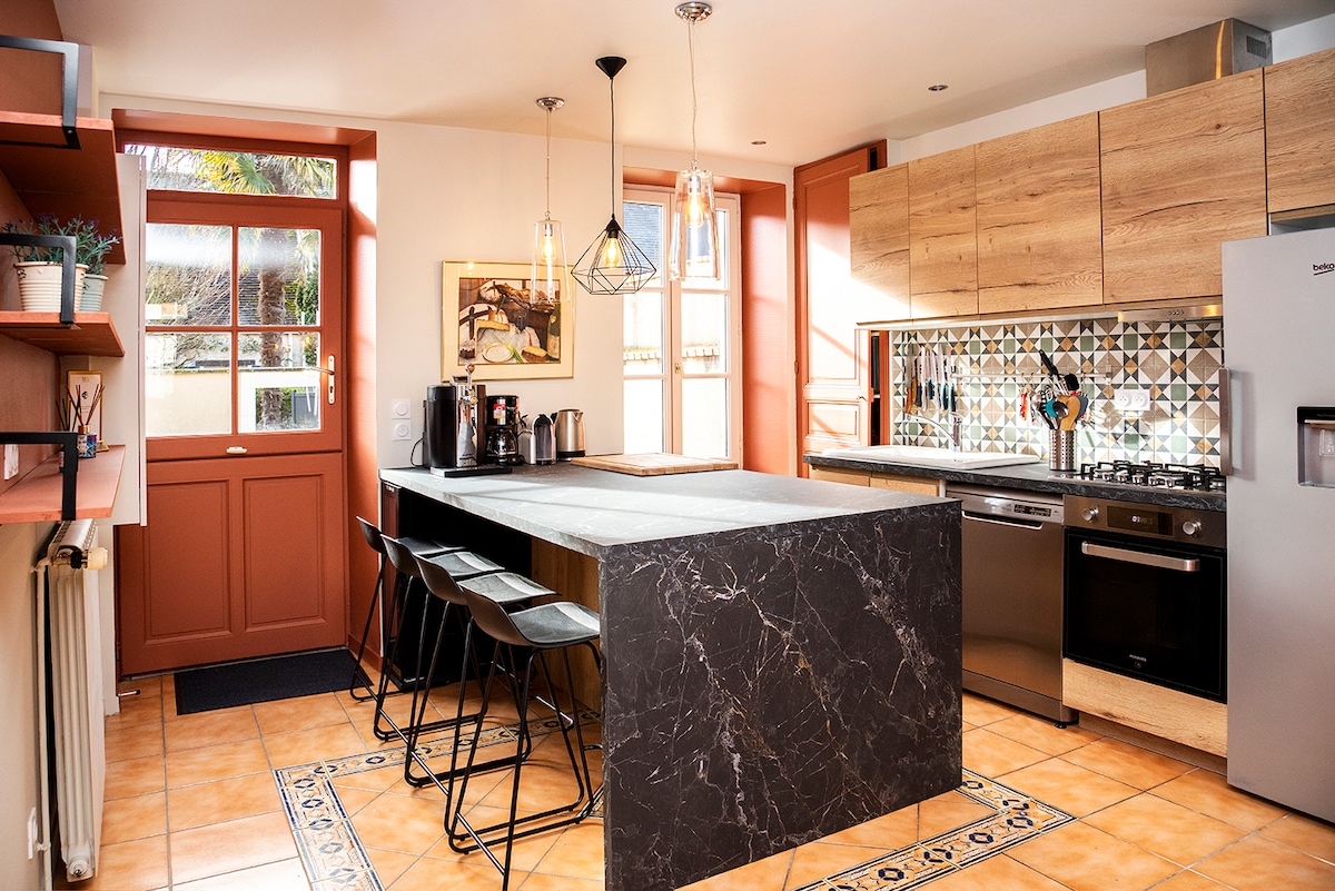 A modern kitchen is featured with a central island covered in dark stone. Wooden cabinetry provides a warm contrast, while patterned tiles decorate the backsplash. Natural light enters through large windows, and a dining area with high-backed stools enhances the space.