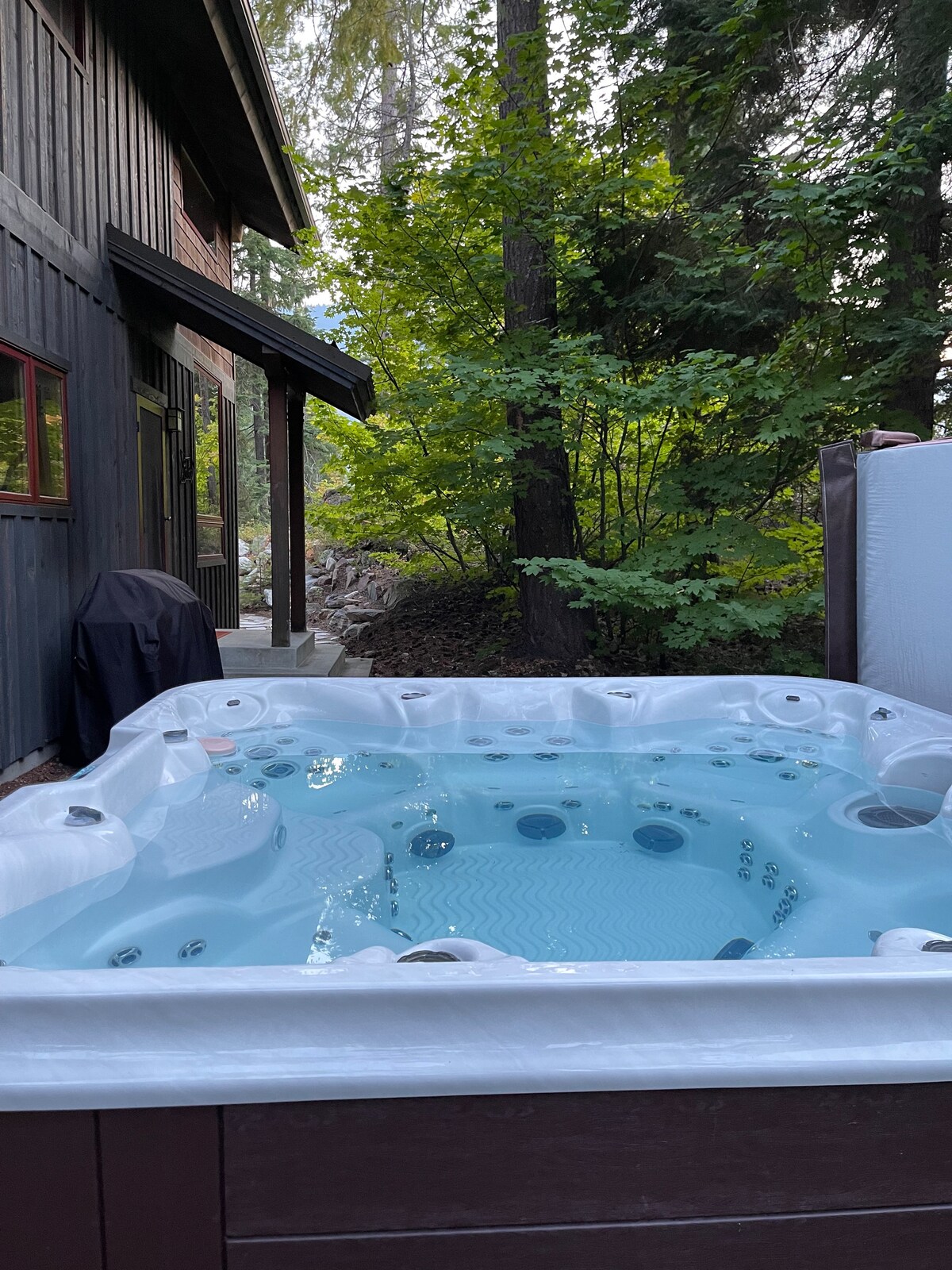 A hot tub is situated in a serene outdoor setting, surrounded by lush green trees. The clear water is inviting, and the arrangement creates a peaceful atmosphere for relaxation.