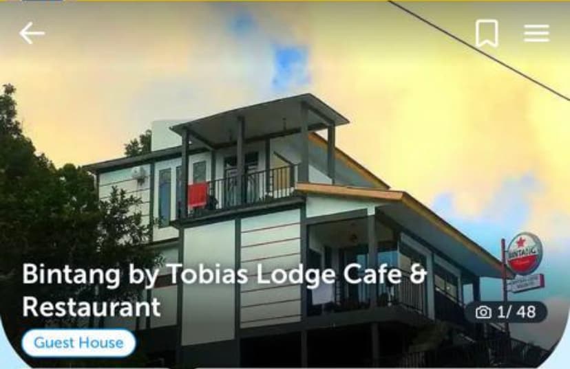The exterior of Bintang by Tobias Lodge Cafe & Restaurant is displayed, featuring a modern three-story building. Large glass windows and balconies provide views of the surroundings. A sign with the lodge's name is visible, alongside greenery that adds to the natural ambiance.
