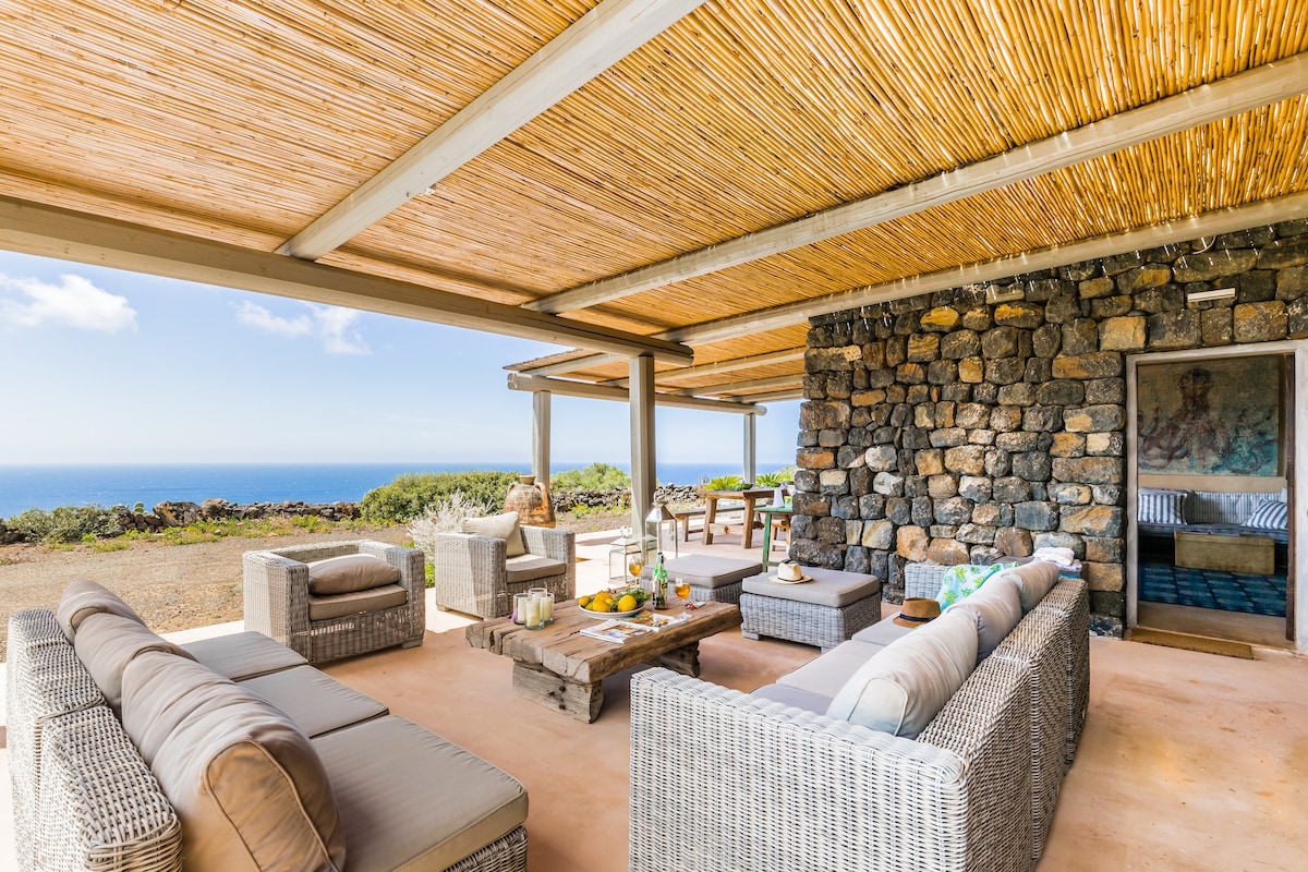 An outdoor lounge area features comfortable woven seating with soft cushions. A wooden coffee table is set with refreshments. The space is shaded by a thatched roof, offering panoramic views of the sea and surrounding landscape.