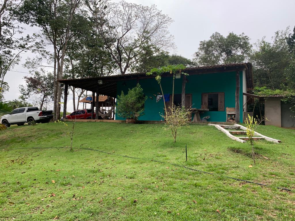 A single-story house with a vibrant green exterior is nestled among trees. A spacious yard with lush grass surrounds the home, which features a covered porch. Several vehicles are parked nearby, indicating accessibility and space for guests.