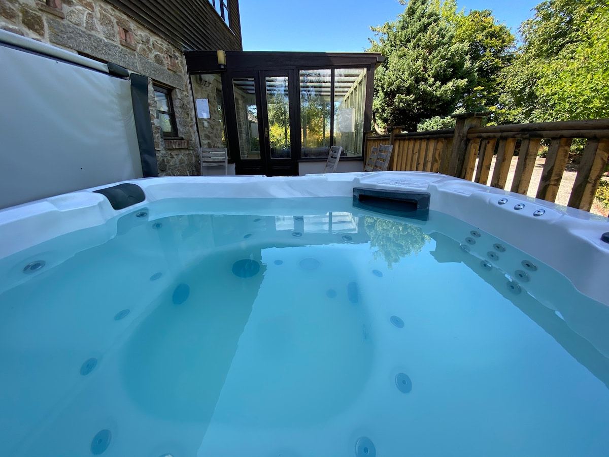 A hot tub with bubbling water is positioned on a wooden deck, inviting relaxation. The backdrop features a glass conservatory with large doors that provide views into the serene garden. Lush greenery surrounds the area, enhancing the tranquil setting.