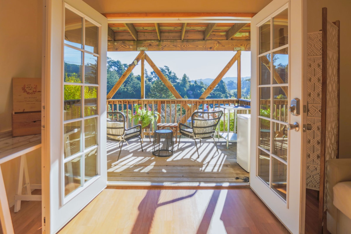 French doors open to a covered deck featuring comfortable seating and a small table. Natural light fills the space, highlighting views of the surrounding hills and greenery. The deck is framed by wooden beams, creating an inviting outdoor area for relaxation.