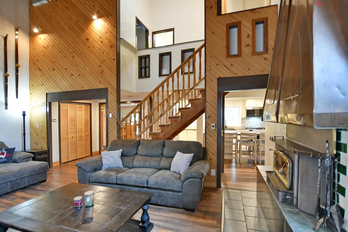 A spacious living area features wooden walls and a high ceiling. A comfortable grey sofa, accented with decorative pillows, is positioned around a dark wooden coffee table. A wood-burning fireplace is visible beside the staircase, with a warm glow from the simple lighting fixtures enhancing the space.
