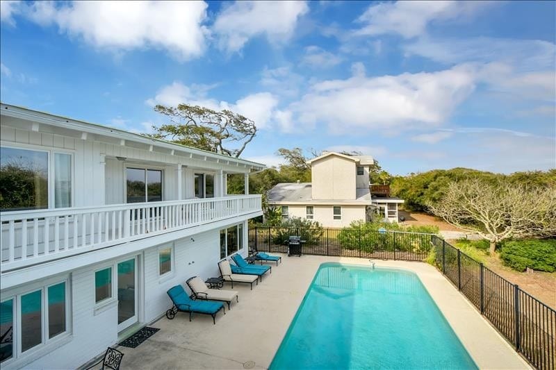 A spacious outdoor area features a large in-ground heated pool surrounded by loungers. The two-story cottage is visible, showcasing multiple windows and a balcony. Lush landscaping and a clear blue sky enhance the inviting atmosphere of the backyard.
