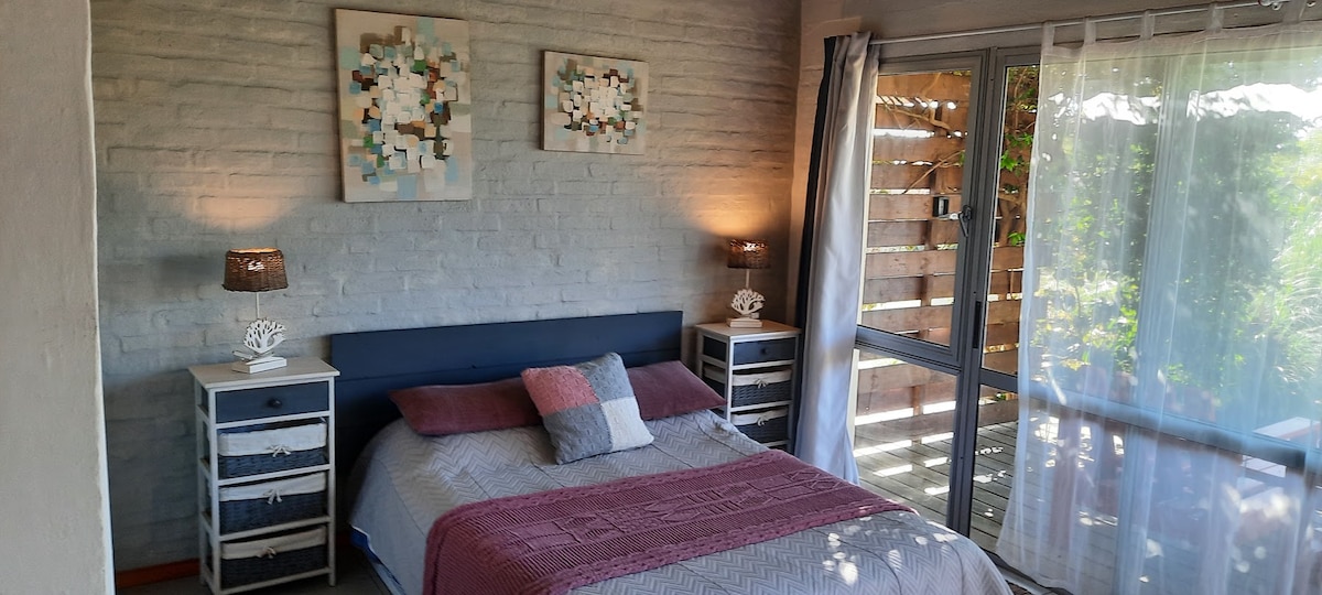 A spacious bedroom features a double bed with decorative pillows and a blanket. Nightstands with lamps are positioned on either side. Large windows allow natural light to enter, complemented by sheer curtains and wooden slats for privacy.