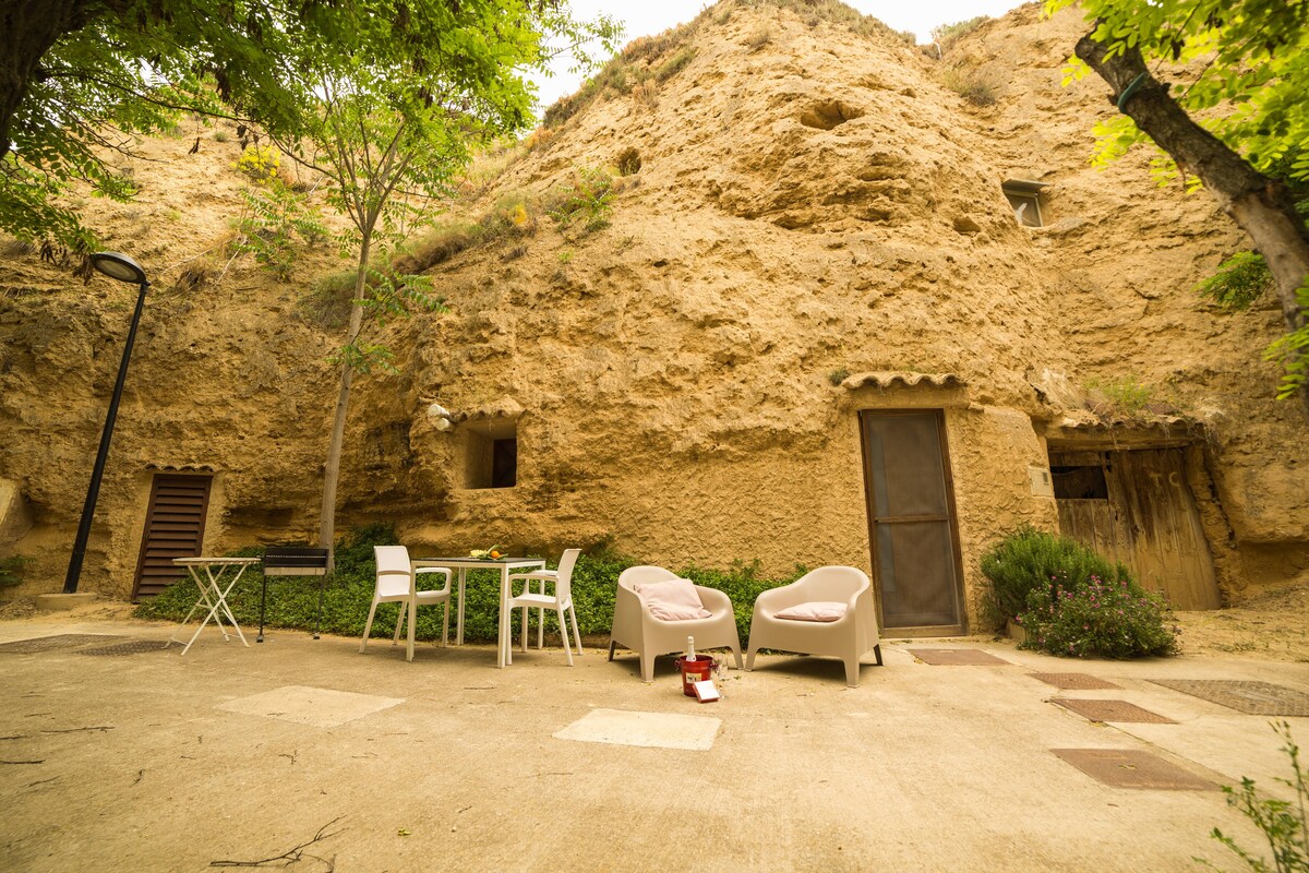 An outdoor area is nestled against a rocky wall, featuring two armchairs and a small dining table with four chairs. A pathway leads to the entrance of the cave. Natural greenery surrounds the space, enhancing the tranquil setting.