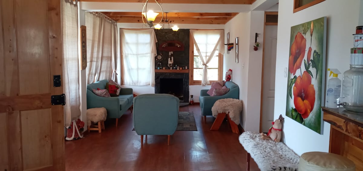 A cozy living area is highlighted by light blue upholstered seating arranged around a central wooden table. A stone fireplace serves as a focal point, and soft curtains adorn the windows, allowing natural light to filter in. A floral artwork adds a touch of color to the wall.
