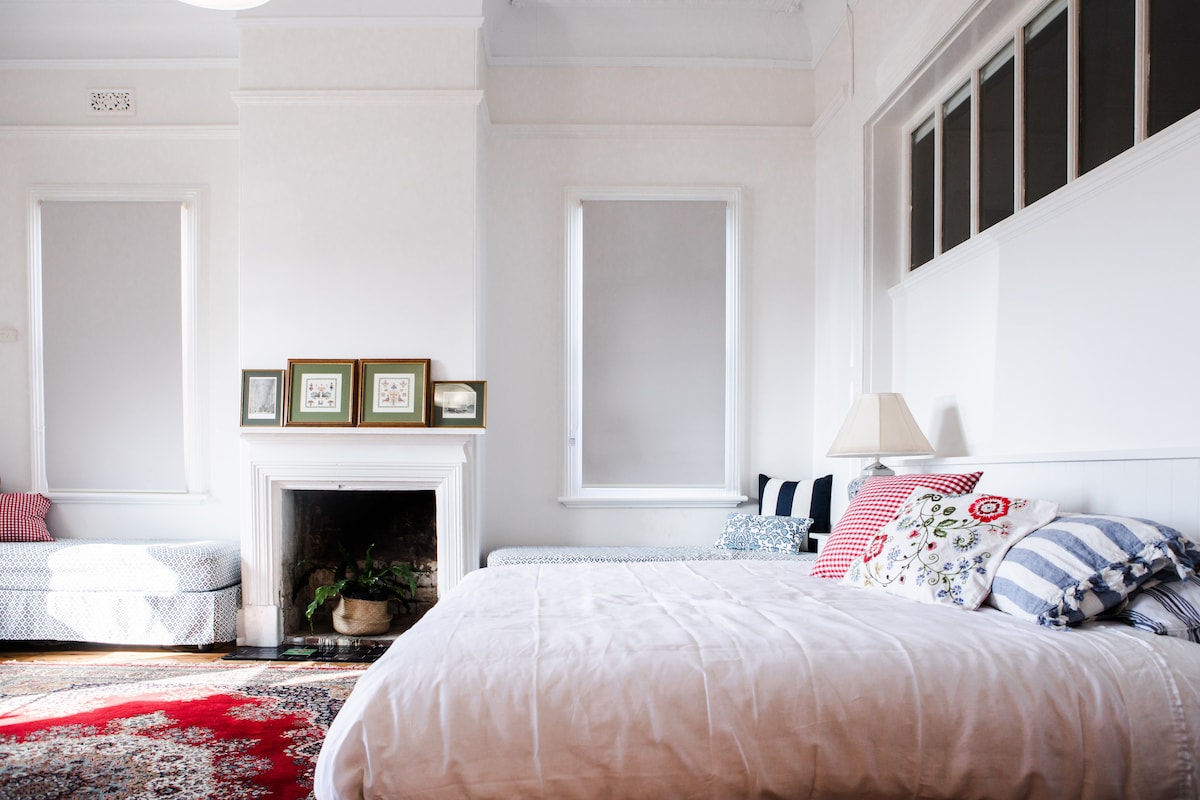 A spacious bedroom is highlighted by a large bed dressed in soft linens. A vintage fireplace is visible, surrounded by framed artwork. Natural light enters through multiple windows with simple shades, while a cozy seating area is provided by a separate bed positioned against the wall.
