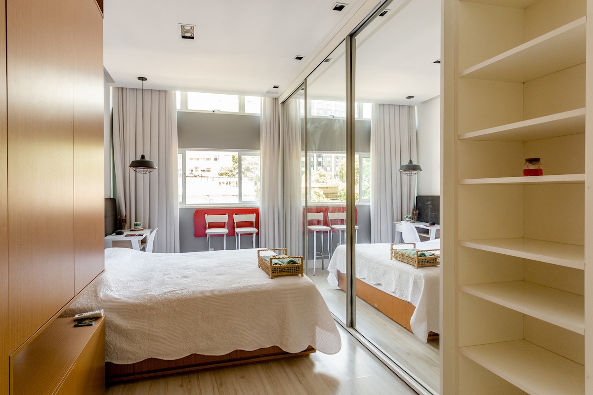 The studio features a spacious sleeping area with a neutral-colored bedspread and large mirrored closets. Natural light filters through the windows, illuminating a small dining space with bar-style seating. A contemporary design is complemented by minimalistic furnishings, ensuring a comfortable atmosphere.