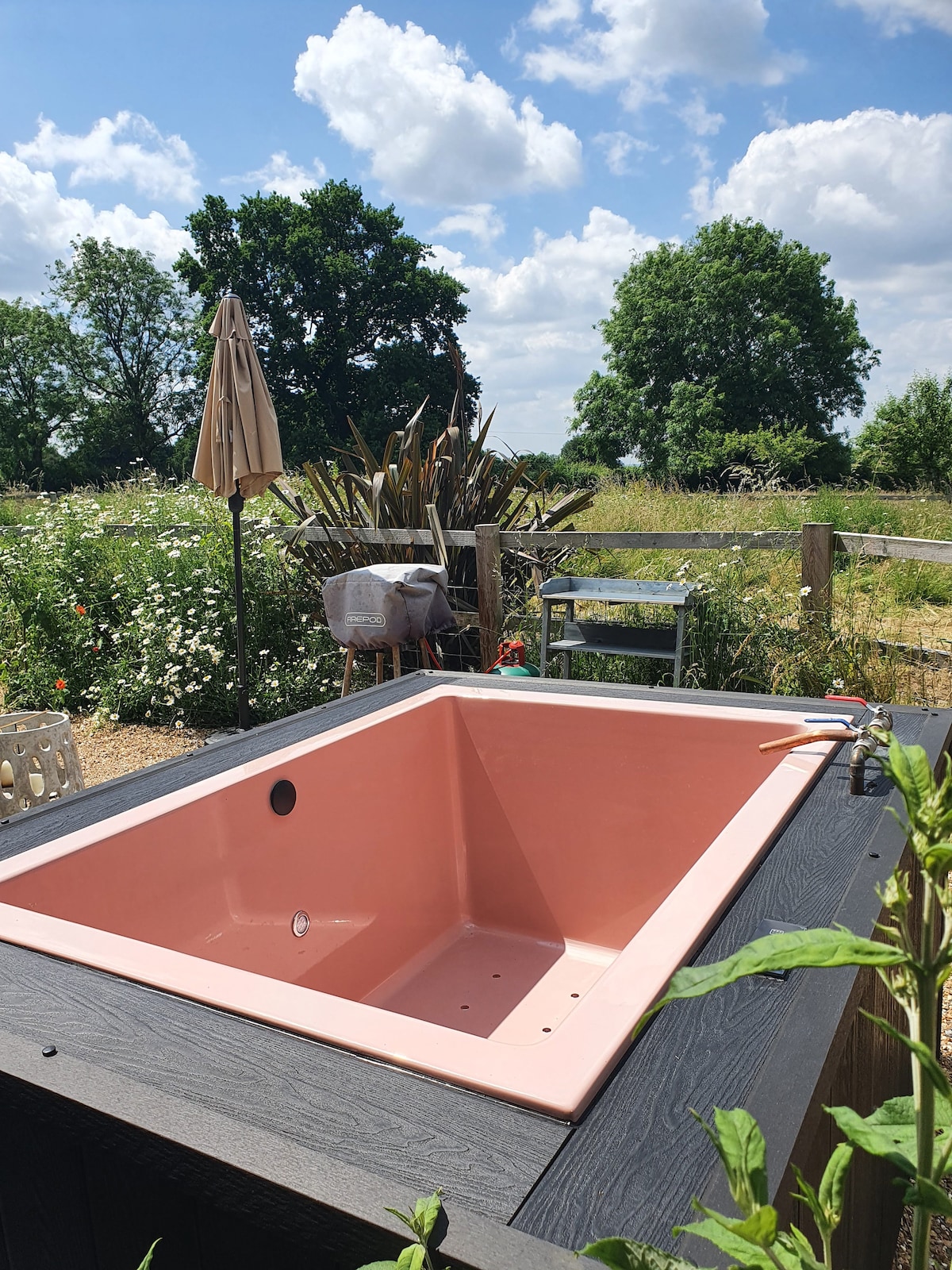 A private Japanese deep soaking tub, featuring a soft pink interior, is positioned outdoors, surrounded by lush greenery and flowering plants. A patio umbrella provides shade nearby, enhancing the serene garden atmosphere.