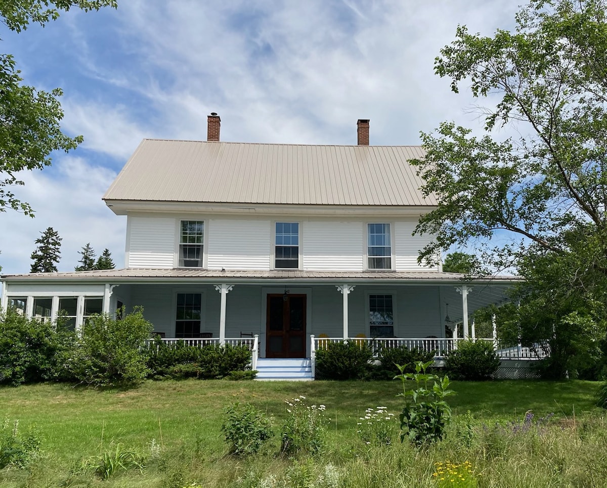 The historic Cow Palace is depicted featuring a wrap-around porch and a spacious lawn. The two-story white structure, topped with a metal roof, is surrounded by lush greenery and flowering plants, providing a welcoming atmosphere that showcases its classic architecture.