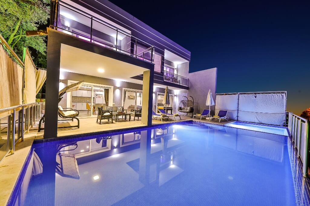An outdoor view of a villa is presented, featuring a spacious swimming pool illuminated at night. The pristine pool reflects the surrounding architecture, while comfortable lounge chairs and a shaded seating area are positioned nearby, offering a cozy atmosphere for relaxation.