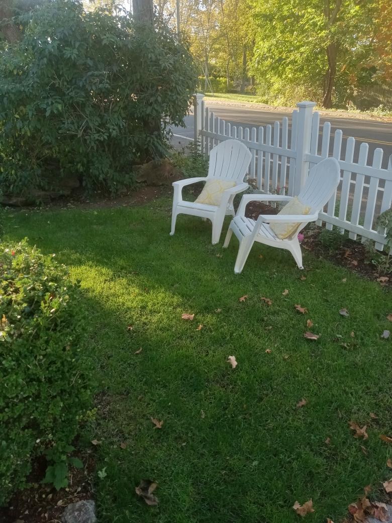 A tranquil outdoor space is depicted, featuring two white lounging chairs positioned on lush green grass. A white picket fence and a flowering shrub provide a serene environment, with filtered sunlight casting gentle shadows across the area.