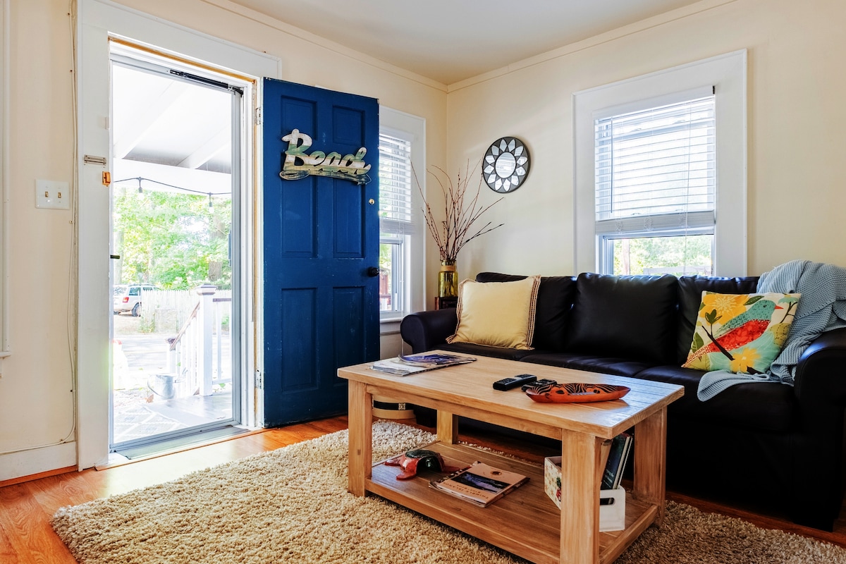 A welcoming entryway features a blue door with a "Read" sign. A dark couch is paired with a colorful accent pillow, and a wooden coffee table displays various items. Natural light streams in through the windows, enhancing the cozy atmosphere.
