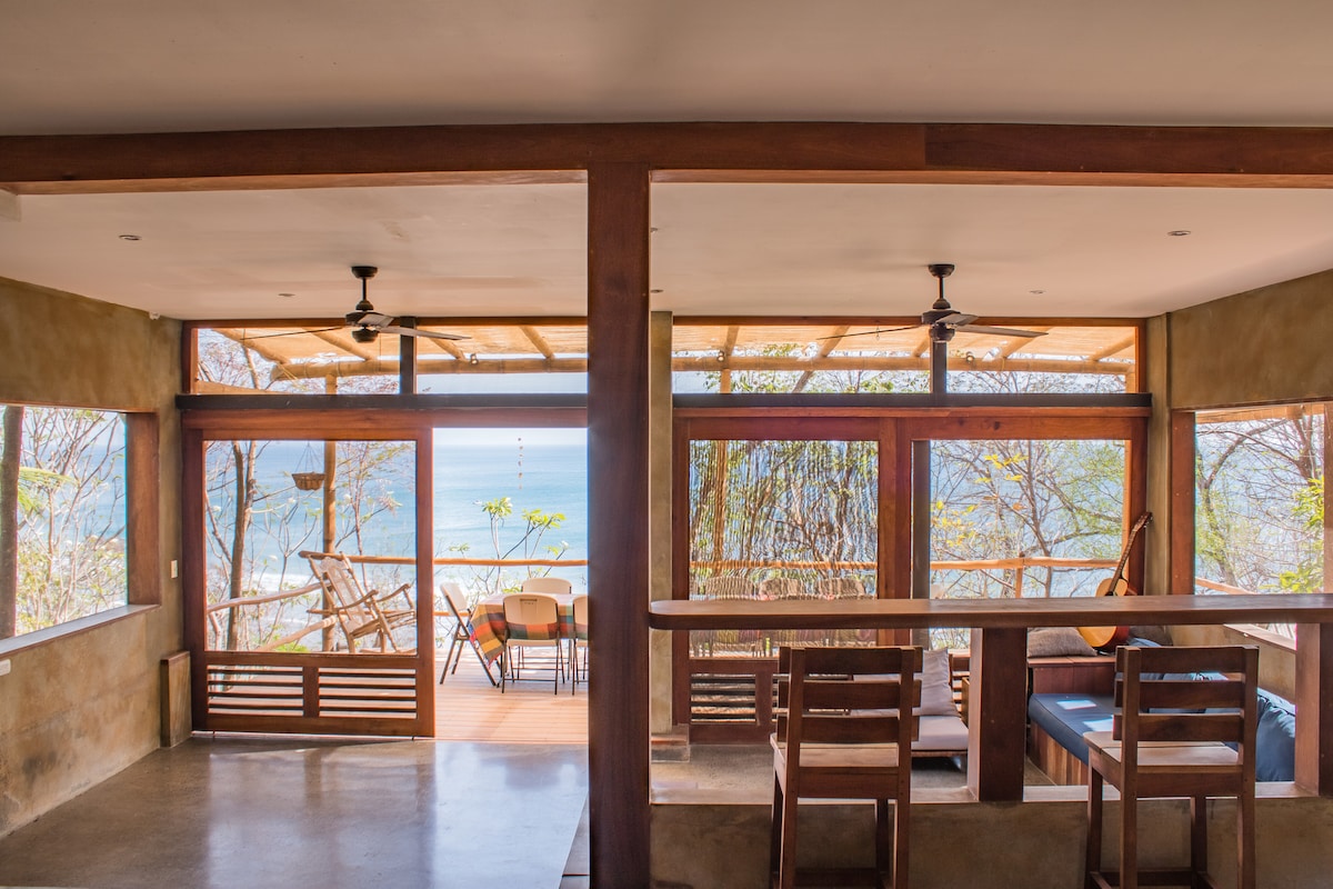 A spacious communal area features large windows that frame a view of the ocean. Wooden furniture, including chairs and a bar counter, is arranged to promote social interaction. Ceiling fans are positioned to enhance airflow, creating a comfortable setting.