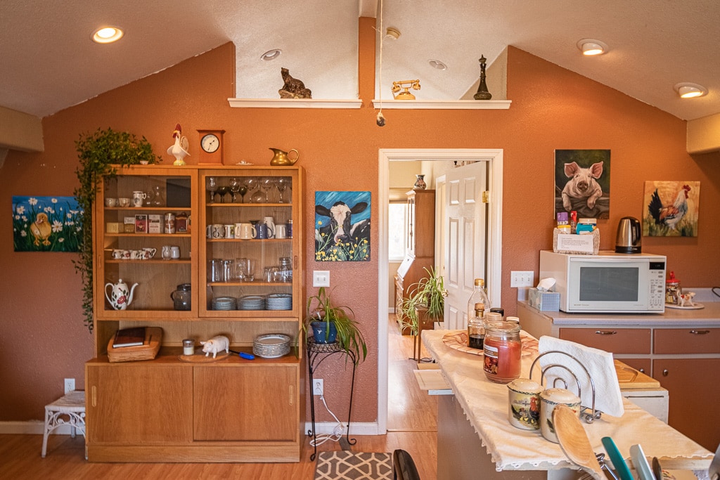 A warm kitchen space features a wooden cabinet displaying various dishes and glassware. An array of colorful artwork adorns the walls, adding character. A microwave occupies the counter, accompanied by a range of jars and utensils. Soft lighting enhances the inviting atmosphere.