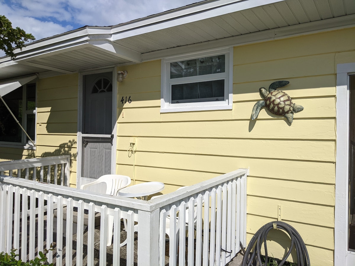 The charming exterior of the Turtle Cottage showcases cheerful yellow siding, complemented by white railings and a small porch area. A decorative turtle motif is mounted on the wall, adding a coastal touch. The front door is framed by a simple overhang, enhancing the inviting entrance.