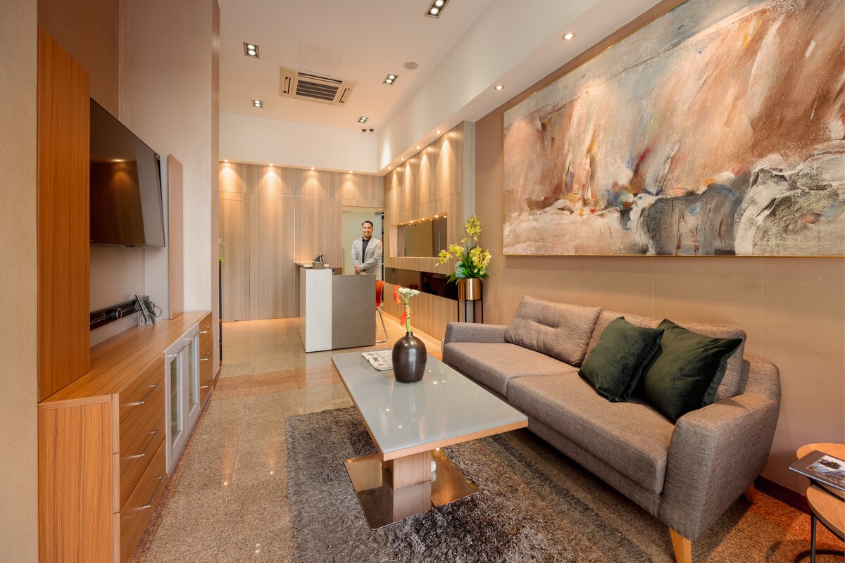 A modern lobby area is depicted with a welcoming atmosphere. A stylish gray sofa is paired with a sleek coffee table at its center, while a large piece of abstract artwork adorns the wall. Reception is visible in the background, with staff ready to assist.
